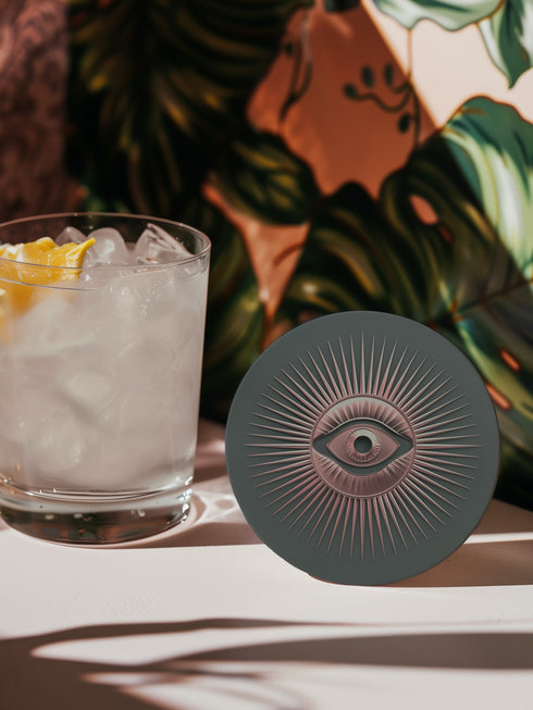 Round ceramic coaster featuring a sage green background with a detailed rose gold Evil Eye symbol in the center, surrounded by radiating metallic rays in a sunburst pattern.